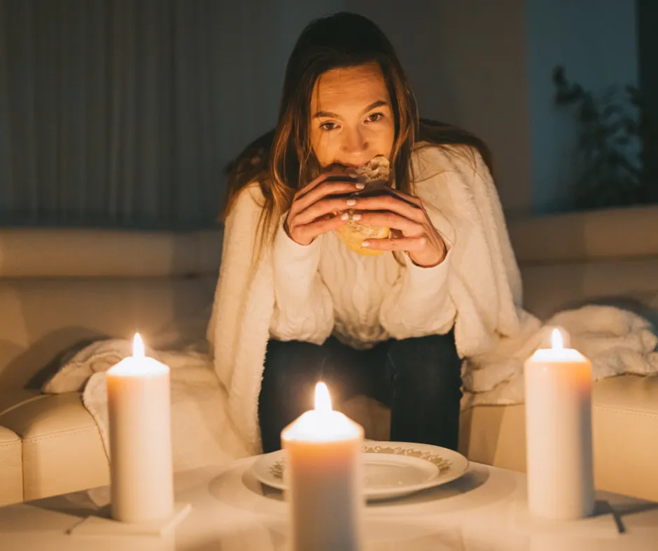 Woman at candlelit table eating burger with three lit candles, relaxed and content in comfortable home setting.