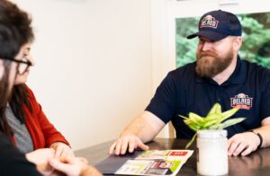 BelRed employee in branded polo shirt and cap discusses service options with female customer at table indoors.