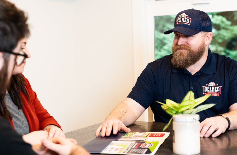 BelRed employee in branded polo shirt and cap discusses service options with female customer at table indoors.