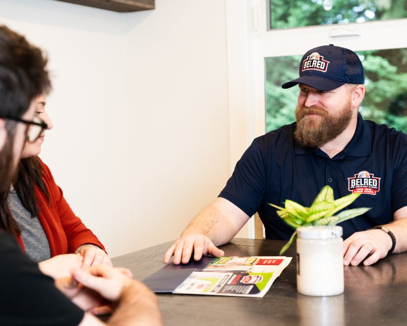 BelRed service professional in branded polo shirt and cap discusses paperwork with female customer at table indoors.