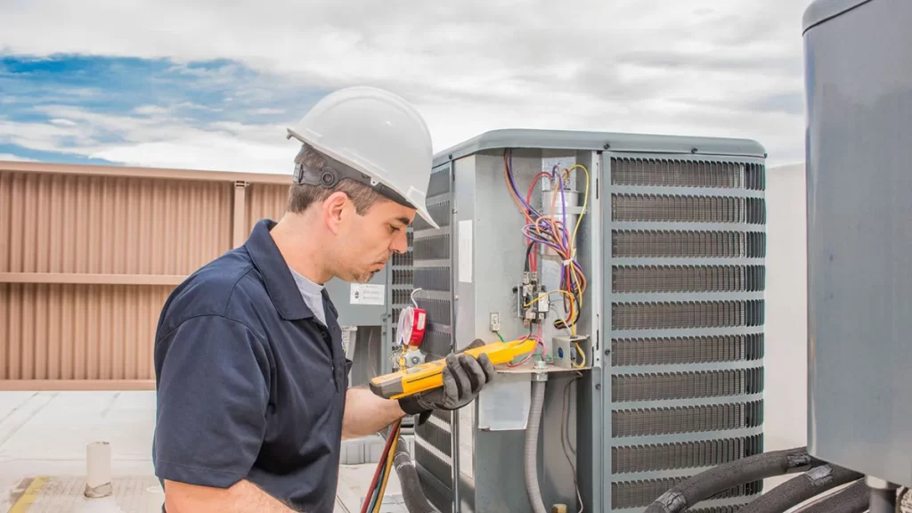 Technician in white hard hat and navy uniform uses diagnostic tool to inspect open air conditioning capacitor unit with visible wiring and components.