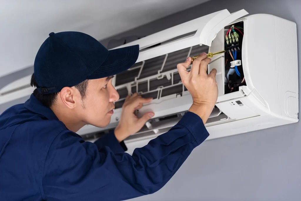 Technician in blue uniform and cap performing maintenance on an air conditioning unit's internal wiring and components.