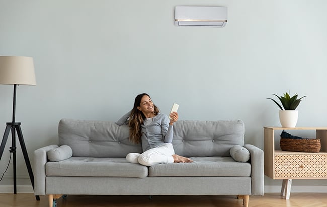 Woman relaxing on gray couch holding remote control with wall-mounted heat pump unit visible above in modern living room.