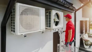 Technician in red shirt and cap standing on ladder while installing outdoor air conditioning unit on white exterior wall.