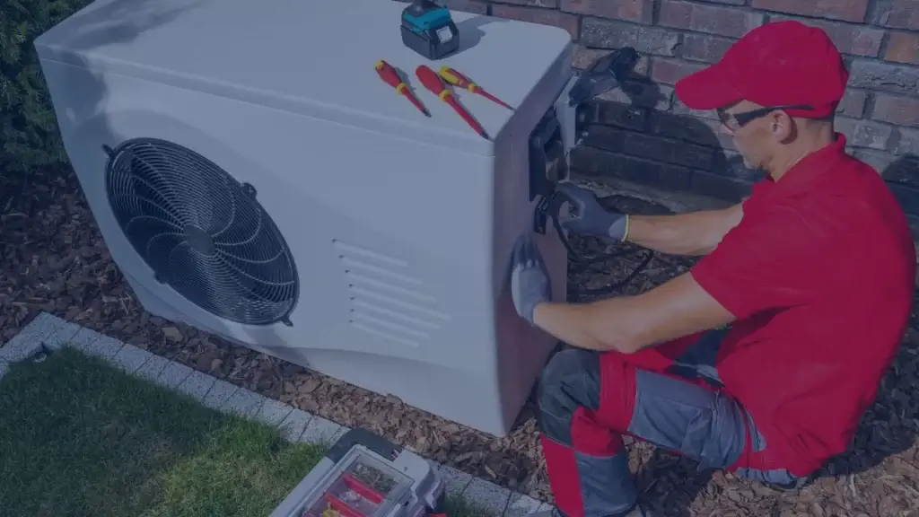 Red-uniformed technician performing maintenance on white outdoor heat pump unit with tools nearby.