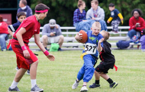 Young football players in blue and black uniforms running with the ball while red team player defends during youth sports event.