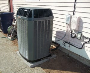 Gray cylindrical heat pump condenser unit installed against white vinyl siding with electrical connections and adjacent equipment visible.