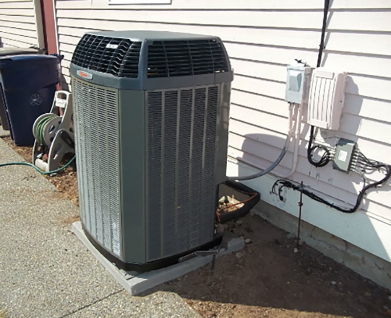 Gray cylindrical heat pump condenser unit installed against white vinyl siding with electrical connections and adjacent equipment visible.